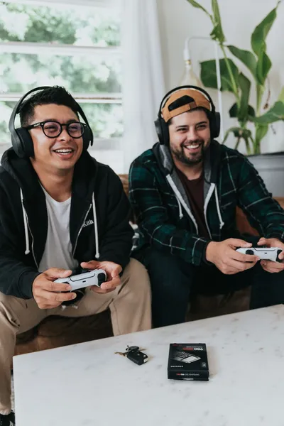 zwei junge Männer sitzen zusammen auf der Couch und spielen mit Controllern ein Videospiel. Sie haben Spaß.