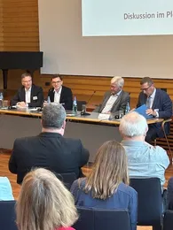 Die Rener*innen des Badischen Dialogs sitzen vor dem Publikum an einem Tisch und diskutieren