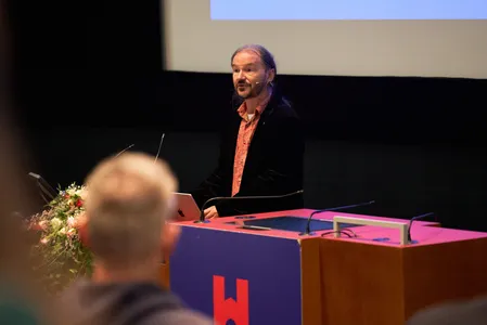 Quelle: Hochschule Offenburg, Hochschule Offenburg Gernot Meier hält einen Vortrag. Ein bärtiger Mann mit langen dunkelgrauen Haaren trägt einen dunklen Anzug