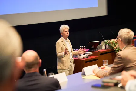 Quelle: Hochschule Offenburg, Hochschule Offenburg Publikum im Vordergrund hört die Rede von Marie-Agnes Strack-Zimmermann. Eine Frau mit kurzem weißen Haar trägt einen sandfarbenen Anzug
